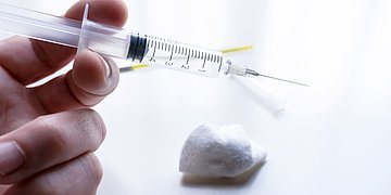 Foto: Eine Hand hält eine Kanüle in der Hand, daneben ein Wattebausch.