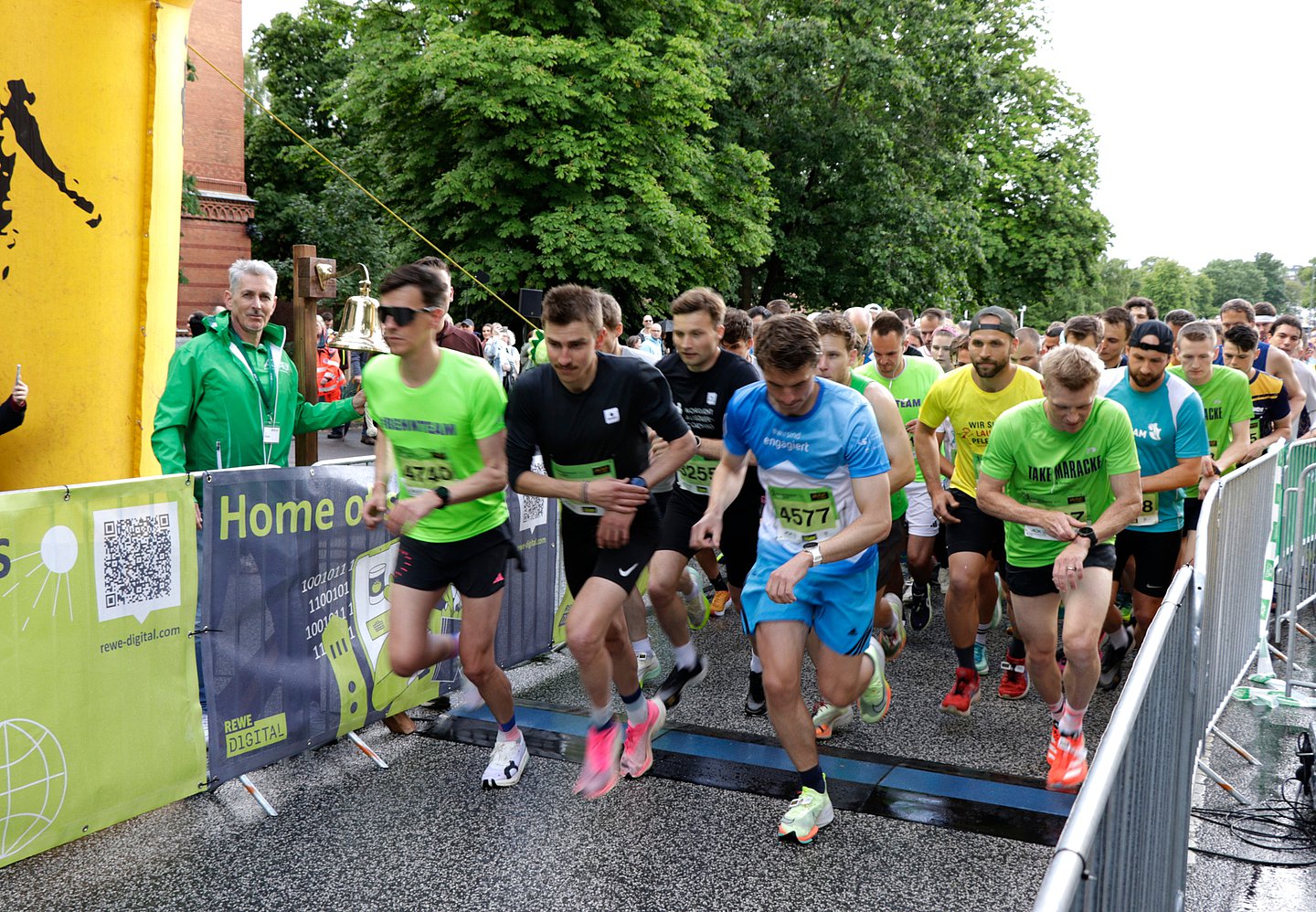 Foto zeigt den Start des AOK-Business.Run Kiel 2024, der mit einer großen Glocke eingeläutet wird, die am linken Bildrand beim Torbogen des Starts zu sehen ist. Das Läuferwelt hat sich gerade in Bewegung gesetzt.