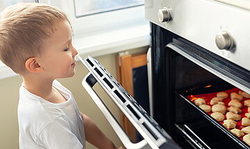 Das Foto zeigt einen kleinen Jungen, der die Backofent&uuml;r &ouml;ffnet, um sich die gebackenen Pl&auml;tzchen anzuschauen.