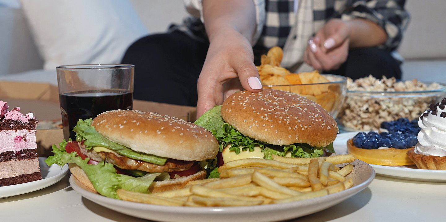 Auf einem Tisch stehen Burger, Pommes, Kuchen, Chips und Popcorn, eine Person greift zum Burger.