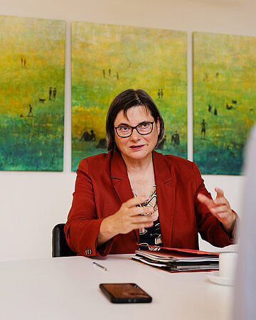 Foto: Christine Fuchsloch, Pr&auml;sidentin des Bundessozialgerichtes (BSG), sitzt an einem Schreibtisch und gestikuliert mit ihren H&auml;nden.