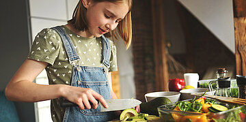 Ein M&auml;dchen in Latzhose und gr&uuml;nem Tshirt mit wei&szlig;en Herzchen steht in der K&uuml;che und schneidet Avocado. Auf der Theke liegen viele Schalen mit Gem&uuml;sesorten sowie &Ouml;lflaschen und Gew&uuml;rze.