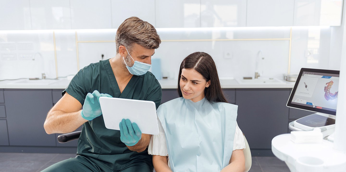 Foto zeigt einen Zahnarzt (links ) im Gespräch mit einer Patientin. Sie sitzt auf dem Zahnarztuhl, während er ihr am Tablet etwas erklärt.