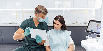 Foto zeigt einen Zahnarzt (links ) im Gespr&auml;ch mit einer Patientin. Sie sitzt auf dem Zahnarztuhl, w&auml;hrend er ihr am Tablet etwas erkl&auml;rt.