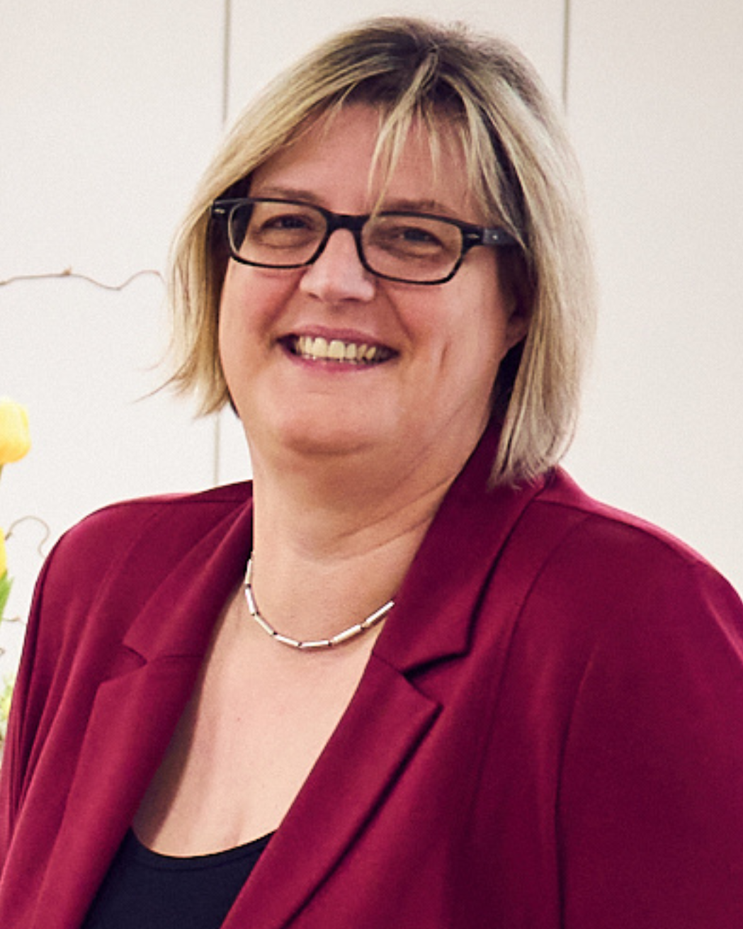 Das Porträtfotozeigt eine blonde Frau mit schulterlangen Haaren einem weinroten Blazer und schwarzem Top darunter
