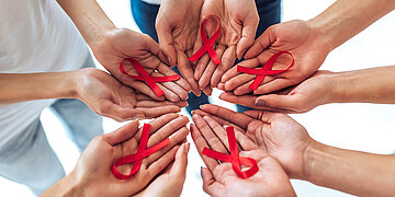 Menschen bilden mit ihren H&auml;nden einen Kreis und haben in den nach oben ge&ouml;ffneten Handfl&auml;chen rote Schleifen als Symbol f&uuml;r Aids.