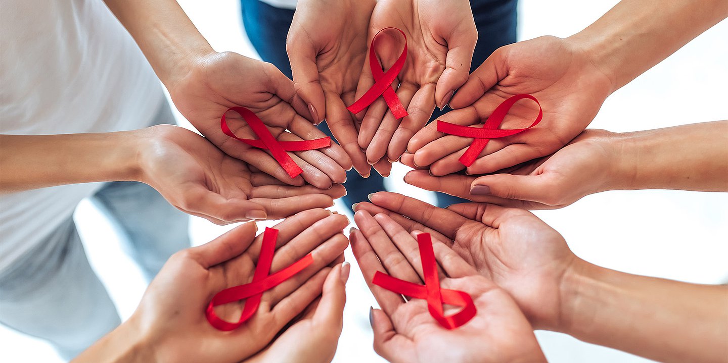 Menschen bilden mit ihren Händen einen Kreis und haben in den nach oben geöffneten Handflächen rote Schleifen als Symbol für Aids.