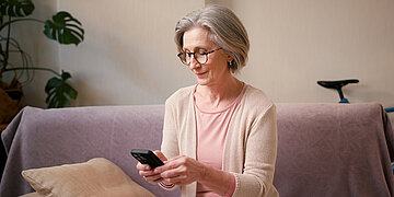 eine &auml;ltere Frau mit grauem Haar sitzt auf einem Sofa und bedient ihr Smartphone.