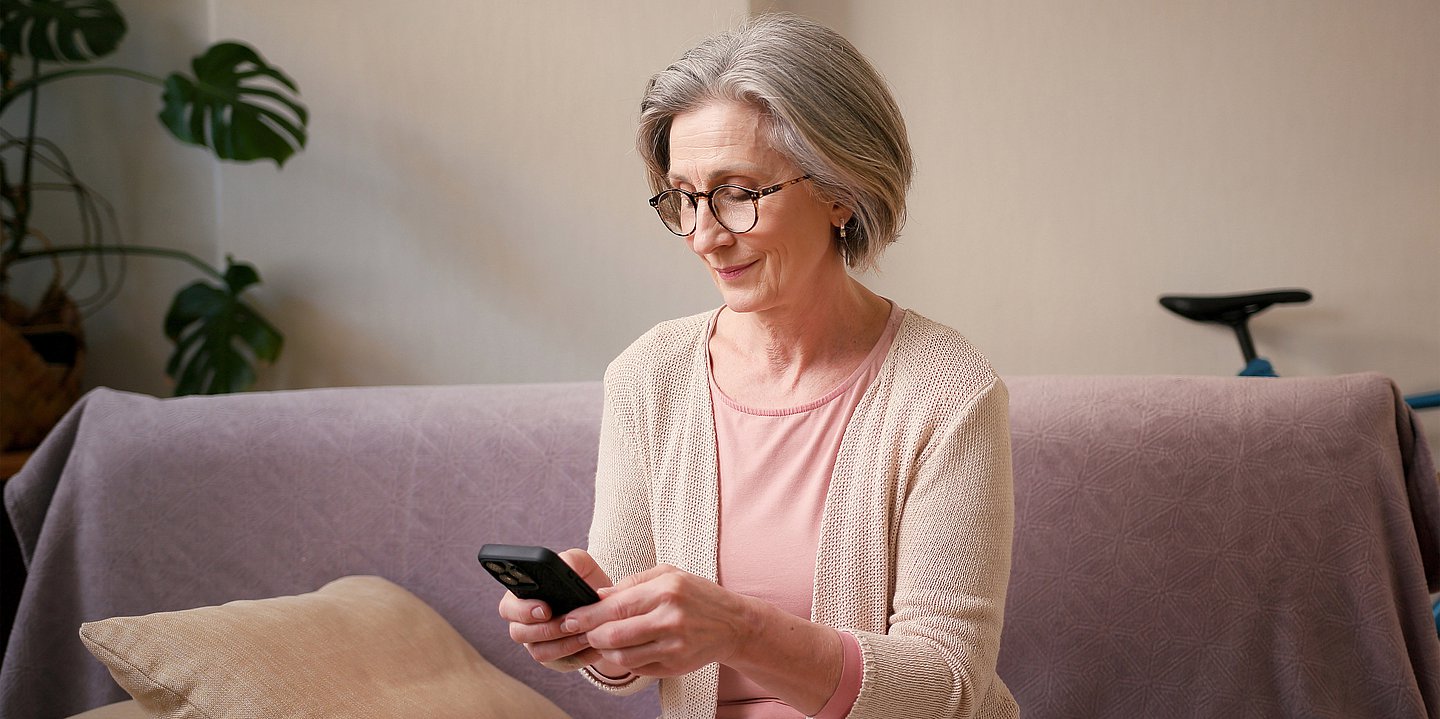 eine ältere Frau mit grauem Haar sitzt auf einem Sofa und bedient ihr Smartphone.