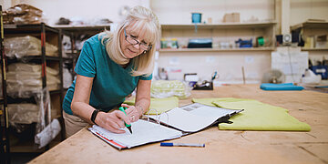 &Auml;ltere Frau arbeitet an einem Tisch und macht Notizen in ein Heft; Regale mit Materialen im Hintergrund.