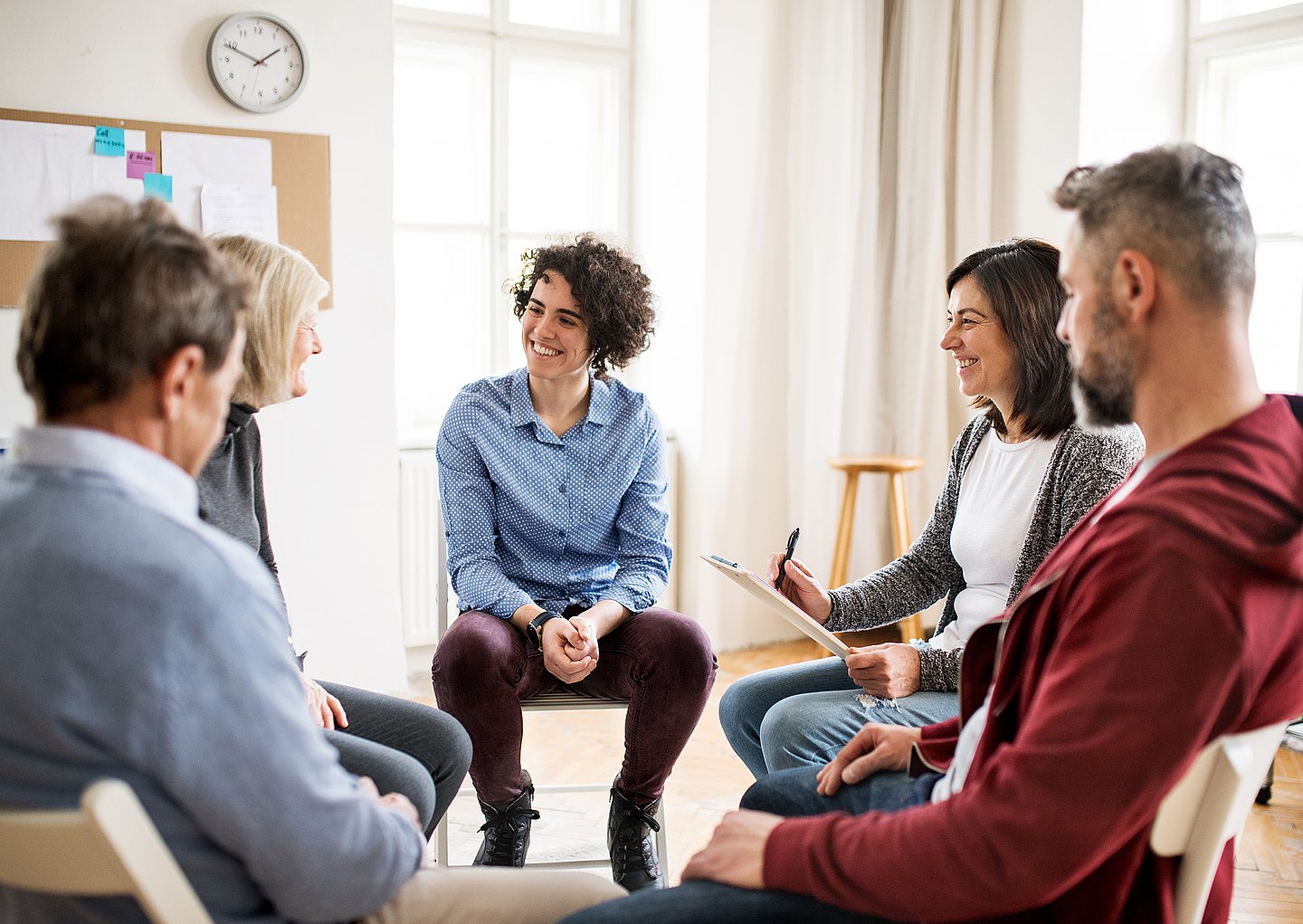 Frauen und Männer sitzen im Stuhlkreis in einem Schulungsraum und tauschen sich aus. Im Hintergrund ist eine Pinnwand mit angehefteten Blättern zu sehen.