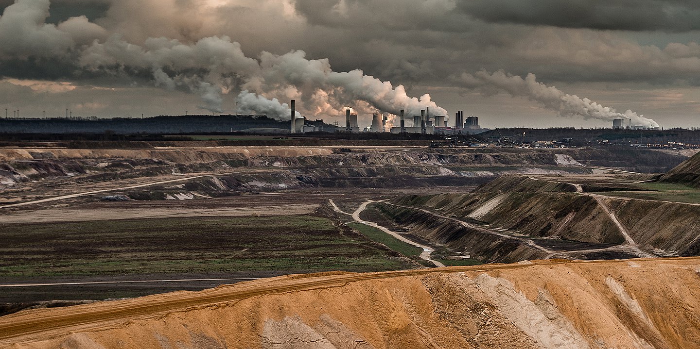 Foto einer ausgebaggerten Landschaft. Im Hintergrund sind rauchende Industrieschlote zu sehen.