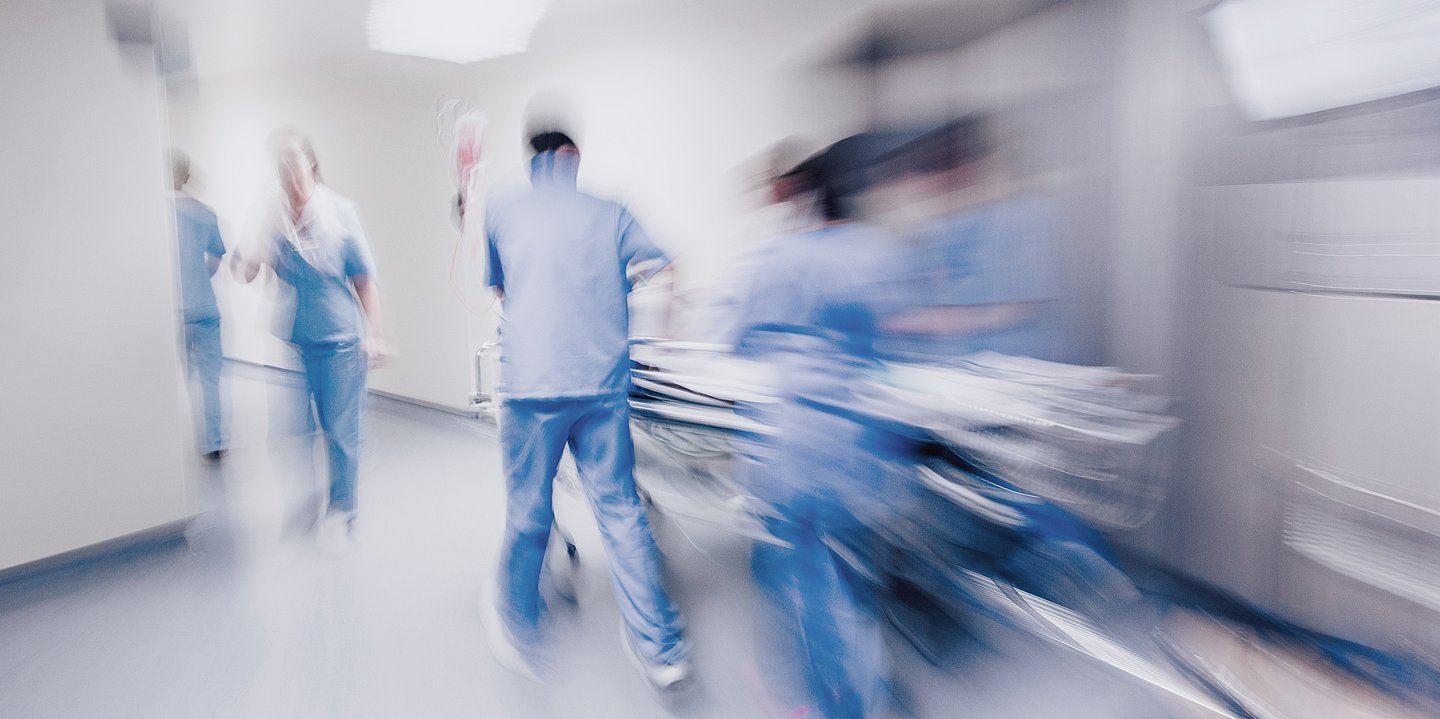 Foto: In einem Klinikflur schiebt Pflegepersonal ein Klinikbett. 