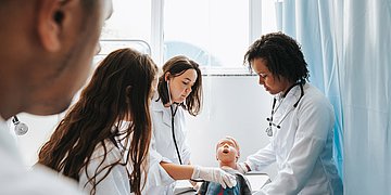 Foto: Zwei Frauen im weißen Kittel üben an einer Kinder-Puppe Pflegeabläufe. Dahinter zwei weitere Kolleg*innen.