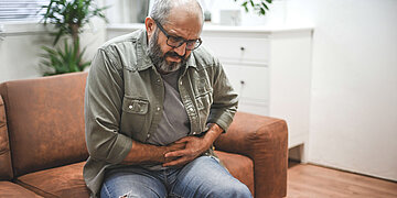 Ein &auml;lterer Herr sitzt auf der Couch, hat ein schmerzverzerrtes Gesicht und fasst sich an den Bauch.