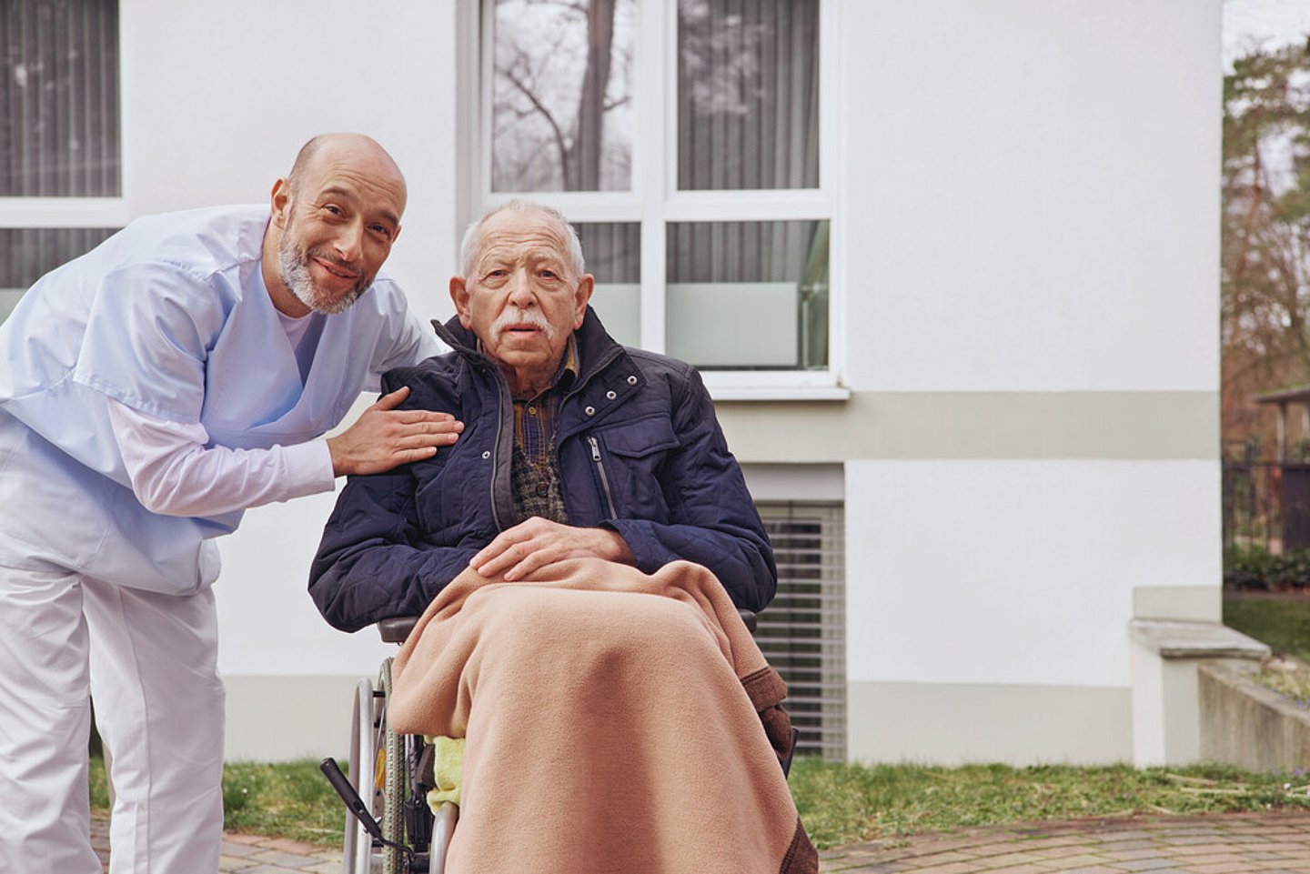 Ein Pfleger mit weißer Hose und blauer Jacke steht neben einem älteren Mann, der mit dunkelblauer Jacke und in eine Decke gehüllt in einem Rollstuhl sitzt. Im Hintergrund befindet sich die Fassade eines modernen Gebäudes, vermutlich ein Pflegeheim.