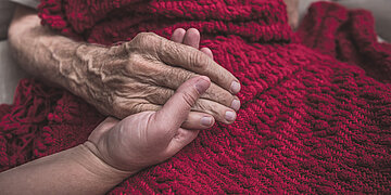 Foto: Eine jungaussehende Hand h&auml;lt eine &auml;ltere aussehende Hand, die H&auml;nde liegen auf einer dunkelroten Wolldecke.