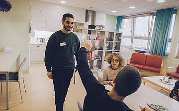 Pfleger Khaled Foto von Pfleger Khaled, wie er freudig auf zwei Kinder zugeht, die in einem Aufenthaltsraum Uno spielen