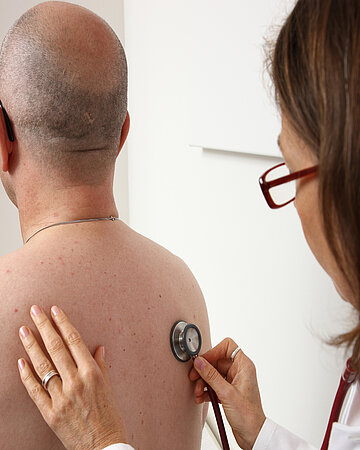 Eine &Auml;rztin untersucht einen Patienten am R&uuml;cken mit ihrem Stethoskop.