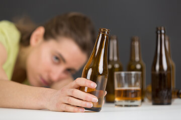 Eine alkoholisierte junge Frau hat den Kopf auf ihren linken Arm abgelegt. Sie schaut auf die leere Flasche, die sie in der rechten Hand ihres ausgestreckten Arms h&auml;lt. In Kopfn&auml;he stehen weitere leere Flaschen Alkohol sowie ein halbvolles Glas.