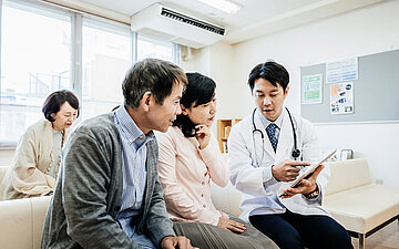 Foto: Ein Arzt sitzt mit einem Tablet in der Hand mit einer Frau und einem Mann zusammen und erkl&auml;rt etwas.