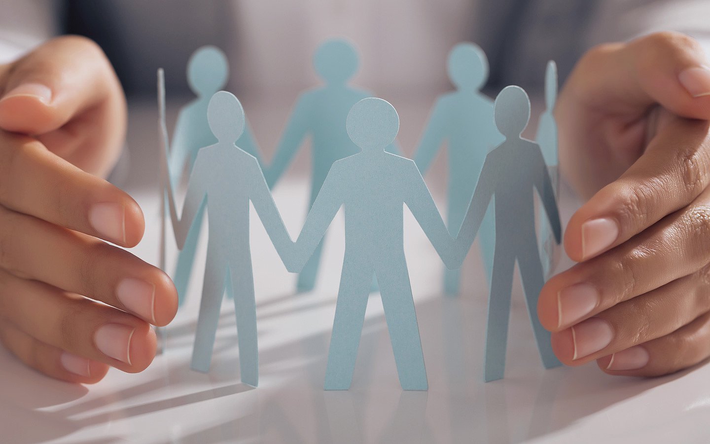 Ein dreidimensionales Schaubild aus einer Gruppe von Menschen in blau zeigt einen Menschenkreis. Zwei Hände sind links und rechts des aufgestellten Menschenkreises schützend aufgestellt.