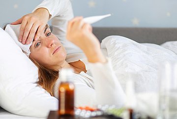 Erkältungswelle in Westfalen-Lippe Das Foto zeigt eine Frau im Bett, die ein Fieberthermometer vor sich abliest.