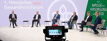 Das Bild zeigt sechs Männer im Halbkreis im TV-Studio. Im Hintergrund ist der weiße Schriftzug "1. Hesseisches Gesundheitsforum" zu lesen. Im Vordergrund steht ein Regie-Monitor