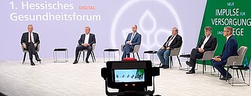 Das Bild zeigt sechs M&auml;nner im Halbkreis im TV-Studio. Im Hintergrund ist der wei&szlig;e Schriftzug "1. Hesseisches Gesundheitsforum" zu lesen. Im Vordergrund steht ein Regie-Monitor