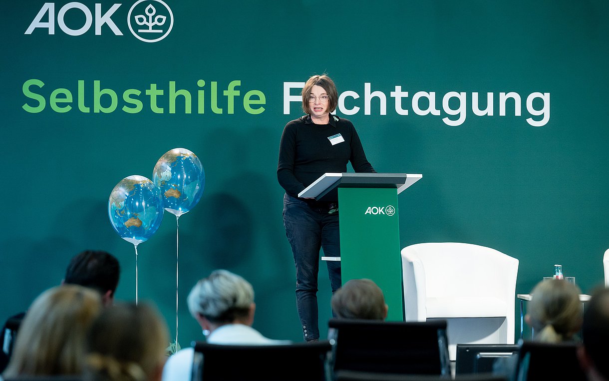 Dr. Annette Scheder steht während ihres Vortrags hinter einem Pult auf einem Podium, im Hintergrund steht an der Wand: Selbsthilfe-Fachtagung