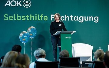 Dr. Annette Scheder steht w&auml;hrend ihres Vortrags hinter einem Pult auf einem Podium, im Hintergrund steht an der Wand: Selbsthilfe-Fachtagung