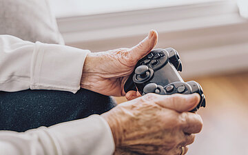 Foto: Zwei H&auml;nde eines &auml;lteren Menschen halten eine Spielkonsole in der Hand.
