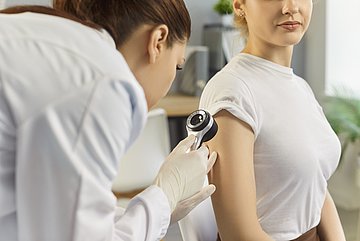 Hautkrebs-Screening in Schleswig-Holstein Eine Ärztin schaut sich mit einer Lupe ein Muttermal am Arm einer jungen Frau an.