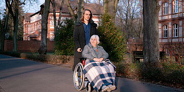 Eine j&uuml;ngere Person schiebt eine &auml;ltere Frau im Rollstuhl auf einem Weg im Freien. Die Seniorin ist mit einer Decke zugedeckt, im Hintergrund stehen B&auml;ume und ein Geb&auml;ude.