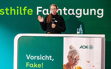 Auf dem Podium steht hinter einem Rednerpult Hanna Leichsenring und spricht in ein Mikrofon. Hinter ihr steht eine Wand mit der Aufschrift "Selbsthilfe-Fachtagung".