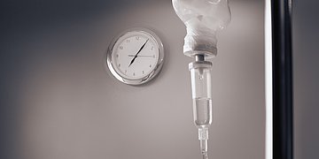 Krankenhausreform – Chronik Bild zeigt im Vordergrund im Anschnitt einen Tropf, im Hintergrund eine runde Wanduhr mit silbernem Rahmen und Ziffernblatt an einer grauen Wand. Die Zahlen sind verschwommen zu erahnen.mit S