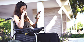 Junge Frai im Rollstuhl, die auf einer sommerlichen Terrasse besorgt in ihre Smartphone blickt