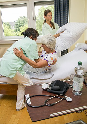 Eine Pflegekraft hilft einer Patientin sich aufzurichten. Währenddessen richtet die andere Pflegekraft das Kissen.