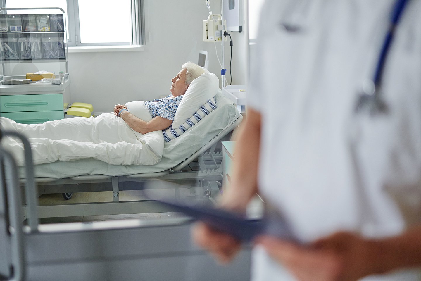 Eine ältere Frau liegt in einem Krankenhausbett. Ein Arzt im Vordergrund schaut auf die Akte.