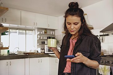 Eine junge Frau steht im rechten Bildausschnitt und hält in der einen Hand einen Becher, in der anderen ihr Smartphone. Sie blickt auf das Display. Im Hintergrund eine große moderne Küche.