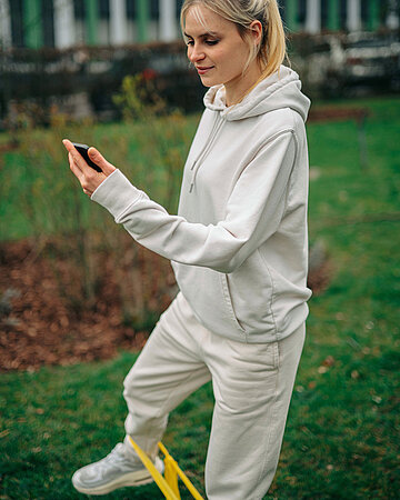 Eine Frau im beigen Trainingsanzug steht auf einer Wiese, macht eine Sport&uuml;bung f&uuml;r die Beine mit einem gelben Trainingsband und schaut dabei in ihr Handy.