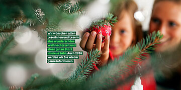 Foto einer l&auml;chelnden Mutter mit Kind hinter einem Weihnachtsbaum. Das Kind h&auml;lt mit einer Hand eine rote Kugel, die am Baum h&auml;ngt.