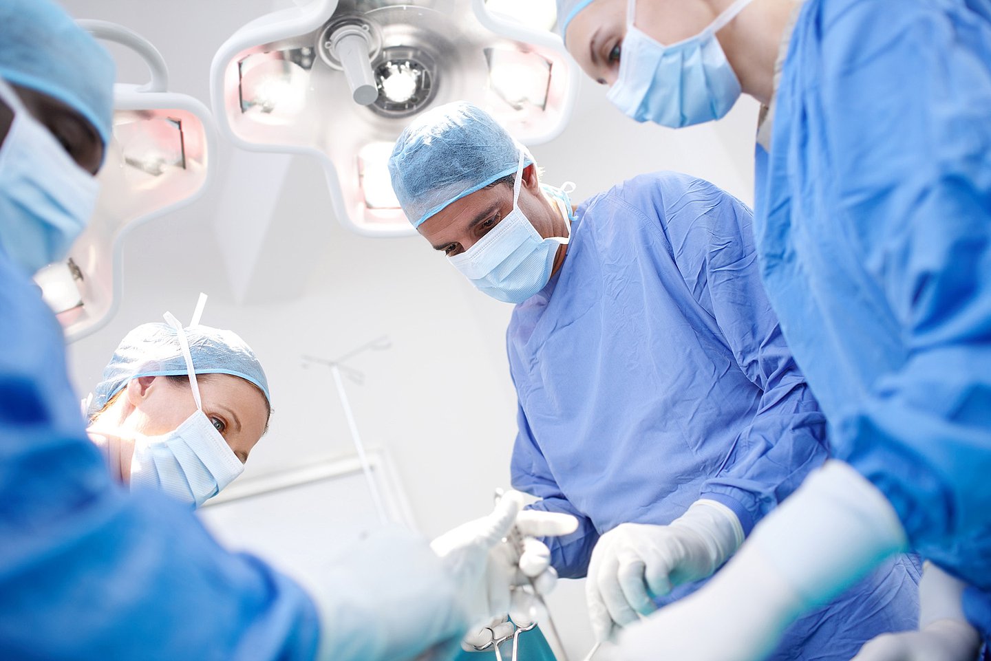 Ein OP-Team steht während einer OP im OP-Saal, alle sind auf den Bereich in der Mitte vor ihnen fokussiert, wo sie mit chirurgischem Werkzeug hantieren.