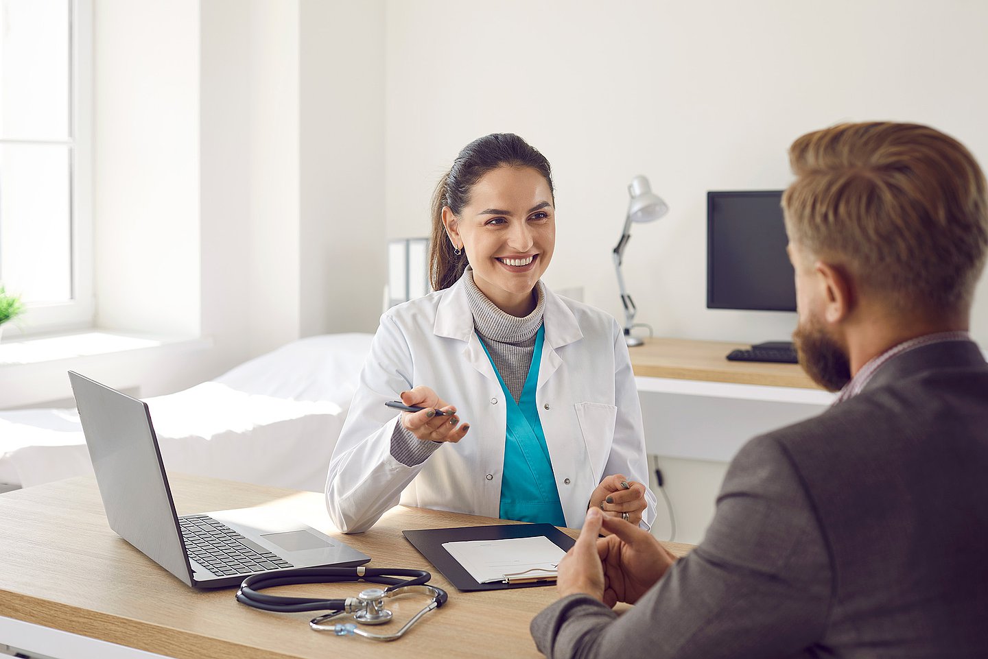 In ihrem Sprechzimmer ist eine Ärztin mit einem Patienten im Gespräch. Vor ihr auf dem Schreibtisch steht ein Laptop, daneben liegt ein Stethoskop.