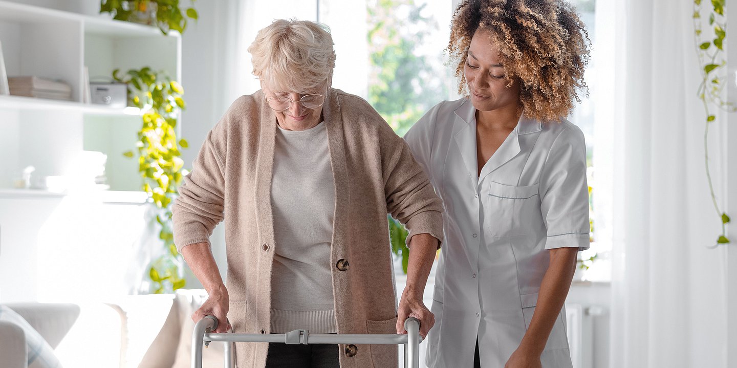 Foto: Eine Pflegekraft hilft einer älteren Frau mit Rollator beim Laufen.