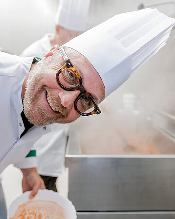 Foto: Sternkoch Maximilian &bdquo;Max&ldquo; Strohe &ndash; mit wei&szlig;er Kochm&uuml;tze &ndash; l&auml;chelt in einer Gro&szlig;k&uuml;che stehend, in die Kamera.