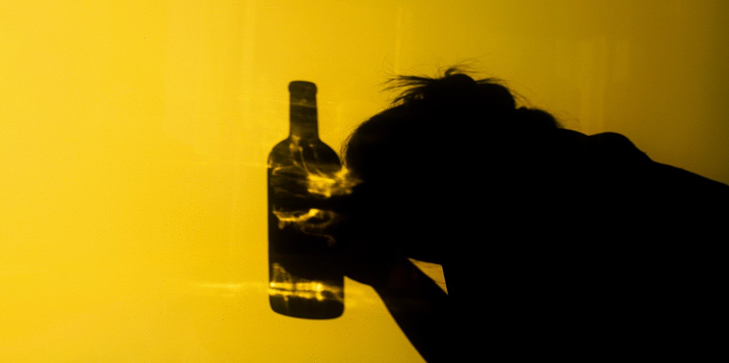 Foto: Schatten einer Frau vor gelbem Hintergrund, die den Kopf auf ihre Hände stützt. Vor ihr – auch als Schatten – eine Flasche.