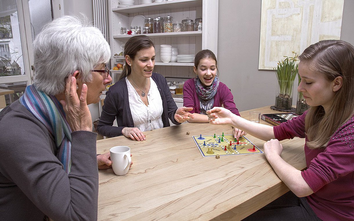 Foto: Kinder und Erwachsene spielen Mensch-ärgere-dich-nicht an einem Tisch. Eine ältere Frau mit Hörgerät beugt sich zu einem Mädchen im Vordergrund des Bildes.