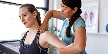 Das Bild zeigt eine Frau in braunem Top, die von einer Physiotherapeutin iin blauem T-Shirt an der linken Schulter behandelt wird. Die Physiotherapeutin steht hinter der Patientin.