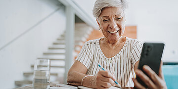 Foto: Eine &auml;ltere Dame h&auml;lt l&auml;cheln ihr Smartphone in der Hand und blickt darauf, w&auml;hrend sie gleichzeitig einen Stift in der Hand h&auml;lt.